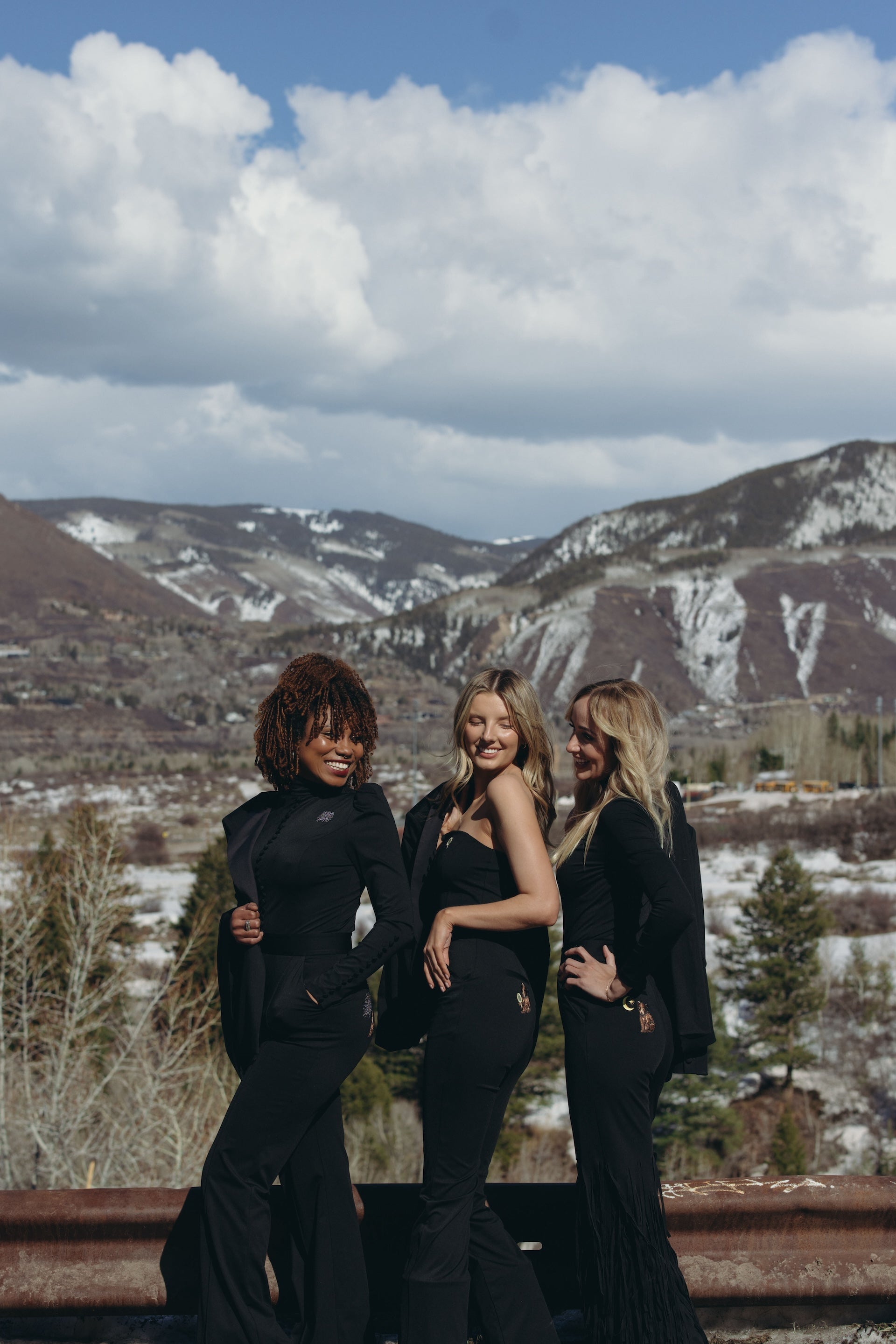 Three women in stylish black Starry Night, Look at Me, and You Hung the Moon jumpsuits pose against a scenic Aspen winter backdrop with mountains and clouds.