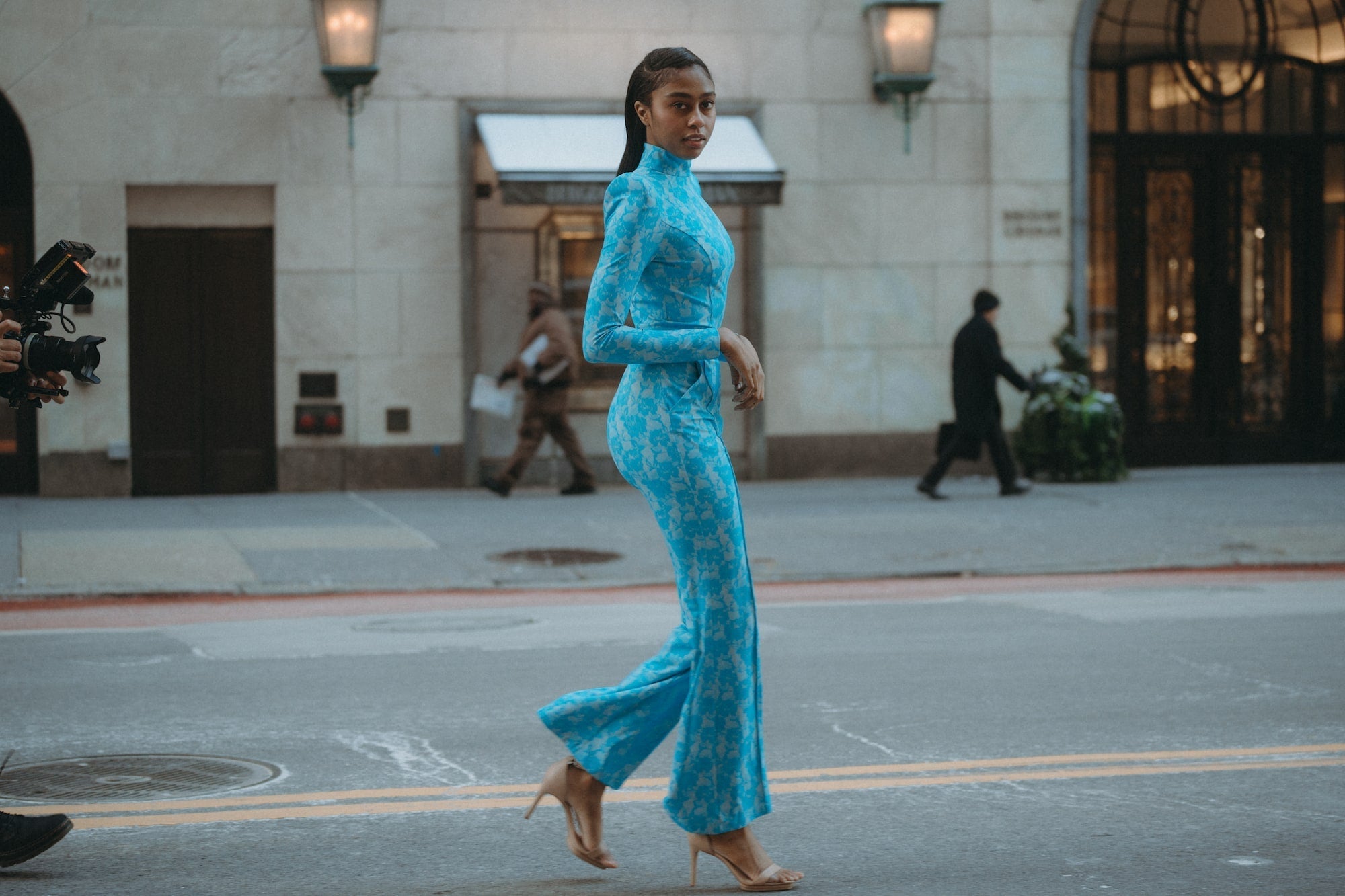 A woman confidently struts in a blue Fellini jumpsuit on a NYC street, showcasing style and elegance during an event.