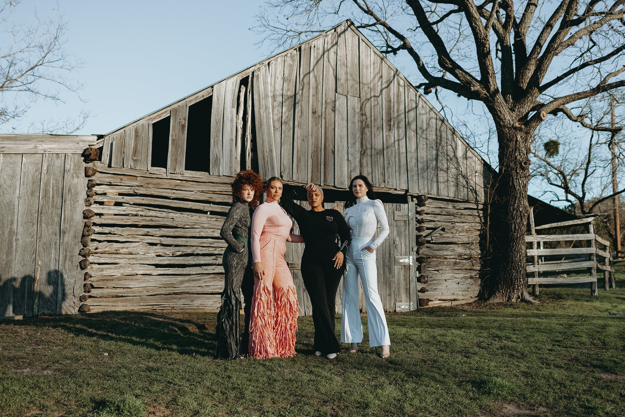 Four women wearing Going Somewhere, Dancing Anyone, Fringy Baby, and I Do jumpsuits pose in front of a rustic barn, showcasing rodeo-inspired fashion choices.