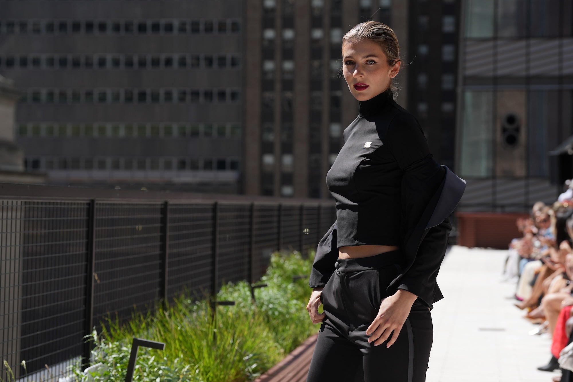 Model showcasing sleek black Tea Time top and pant on a stylish runway, surrounded by an urban backdrop and seated audience.