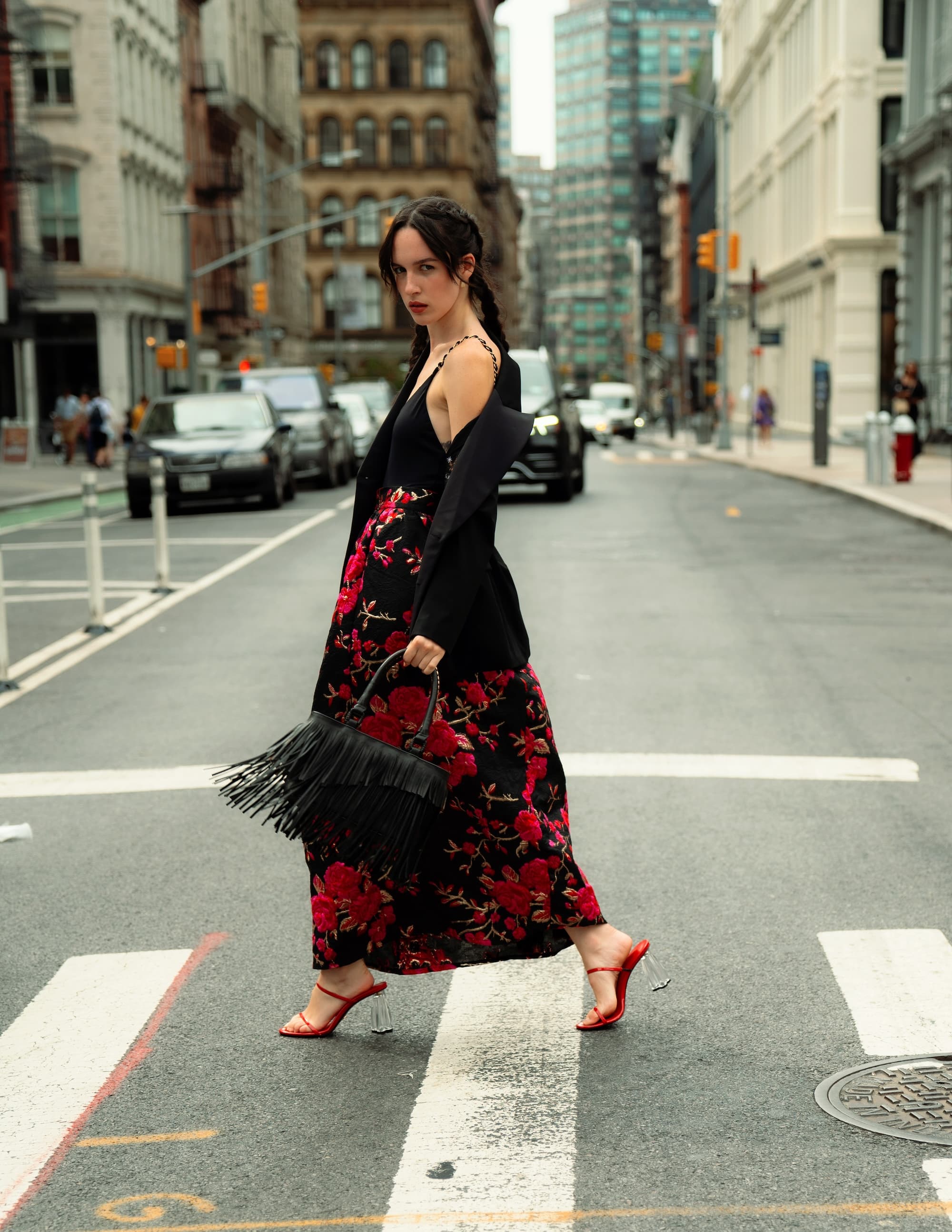 Stylish woman crossing the street in a floral skirt and black cardigan, holding a fringe handbag in an urban setting.