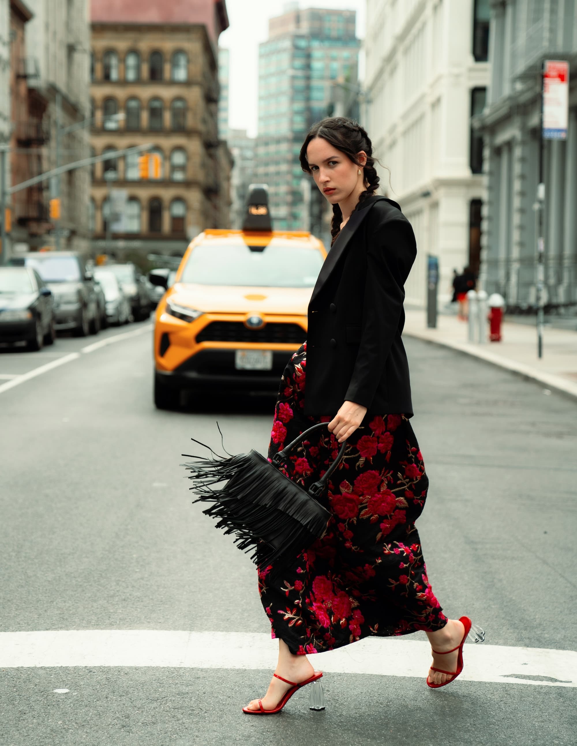 Woman in a floral skirt and blazer walking on the street while holding a stylish black bag, with a taxi in the background.