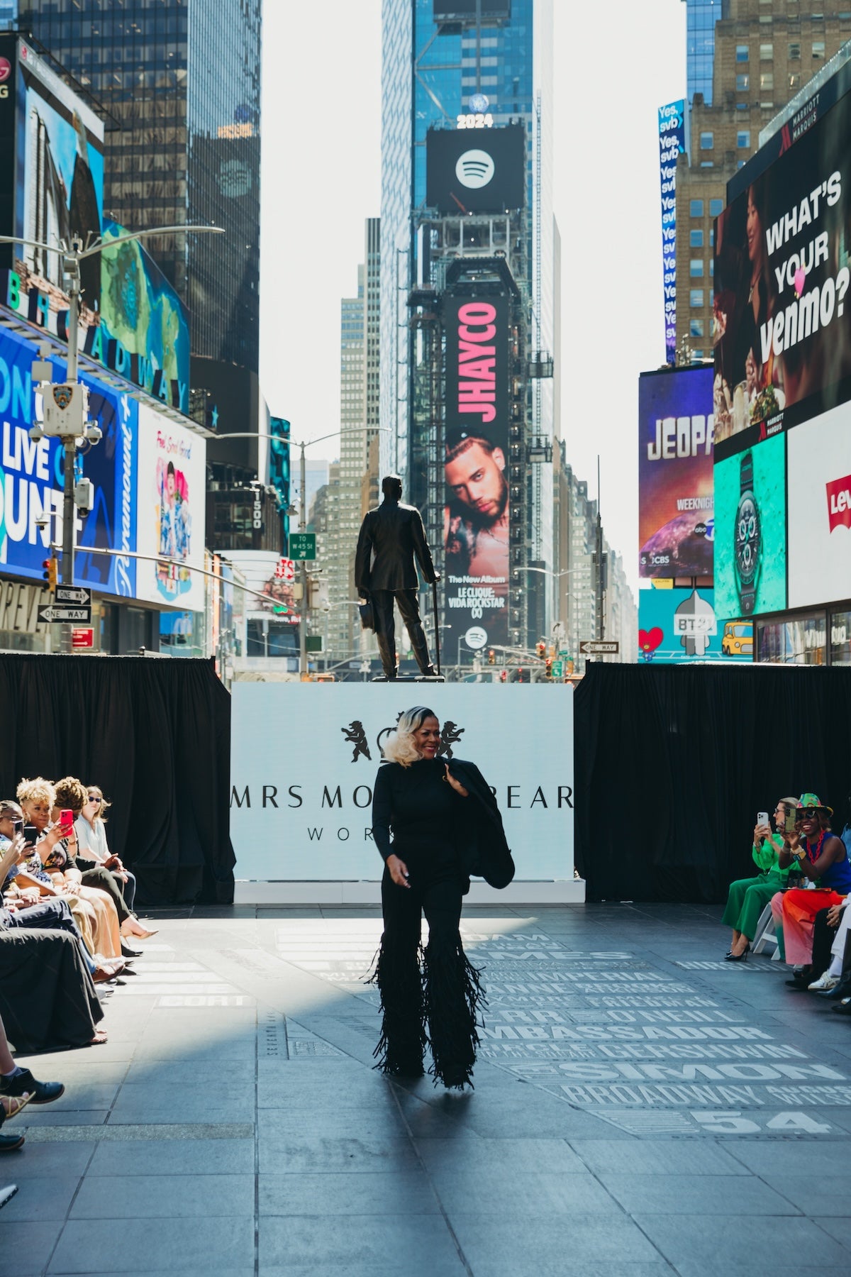 Model walking the runway in Times Square, NYC, showcasing Fringy Baby at fashion show with vibrant billboards in background.