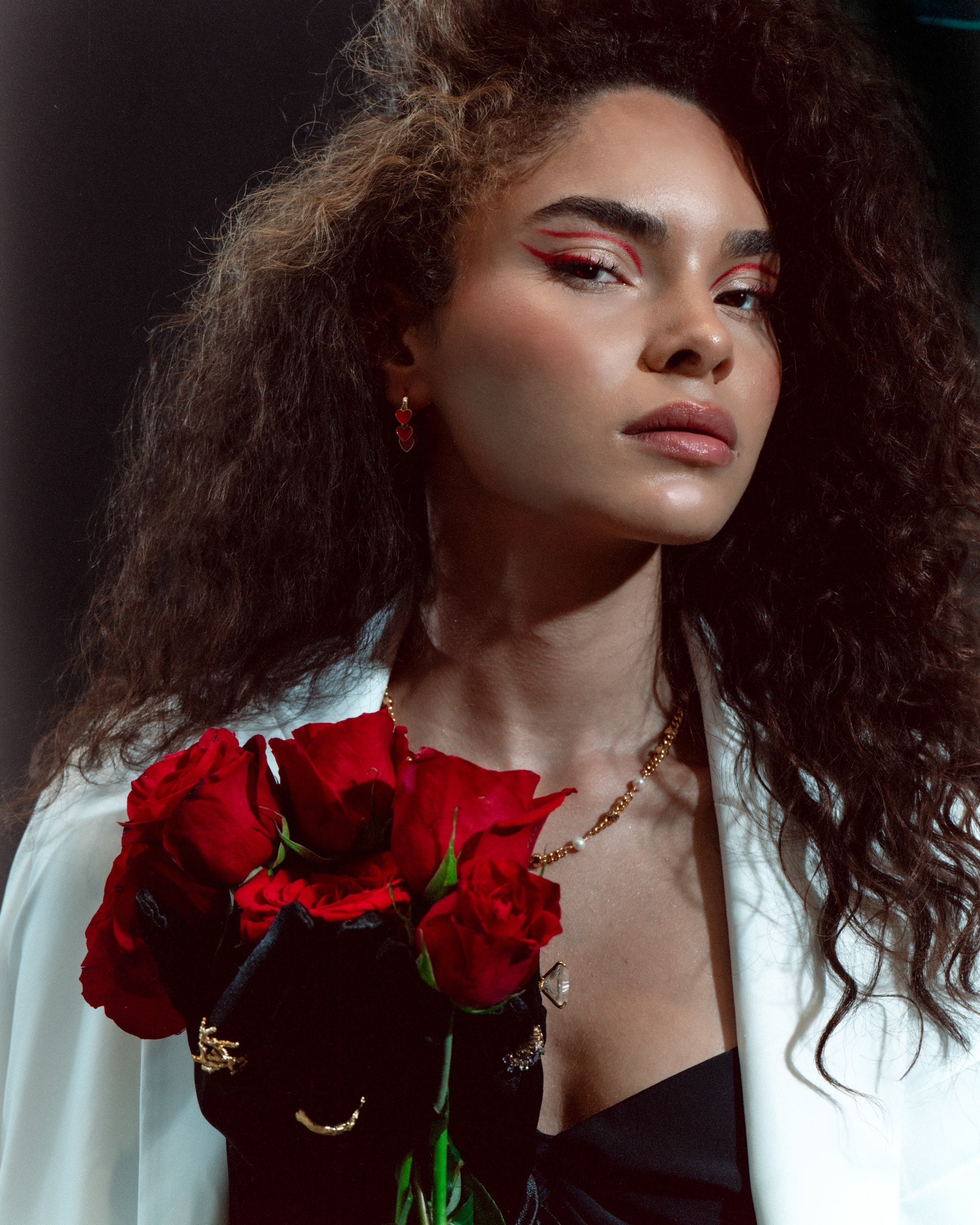 Model with curly hair holding red roses, wearing elegant jewelry and a Meet Me at Noon blazer, exuding confidence and beauty.