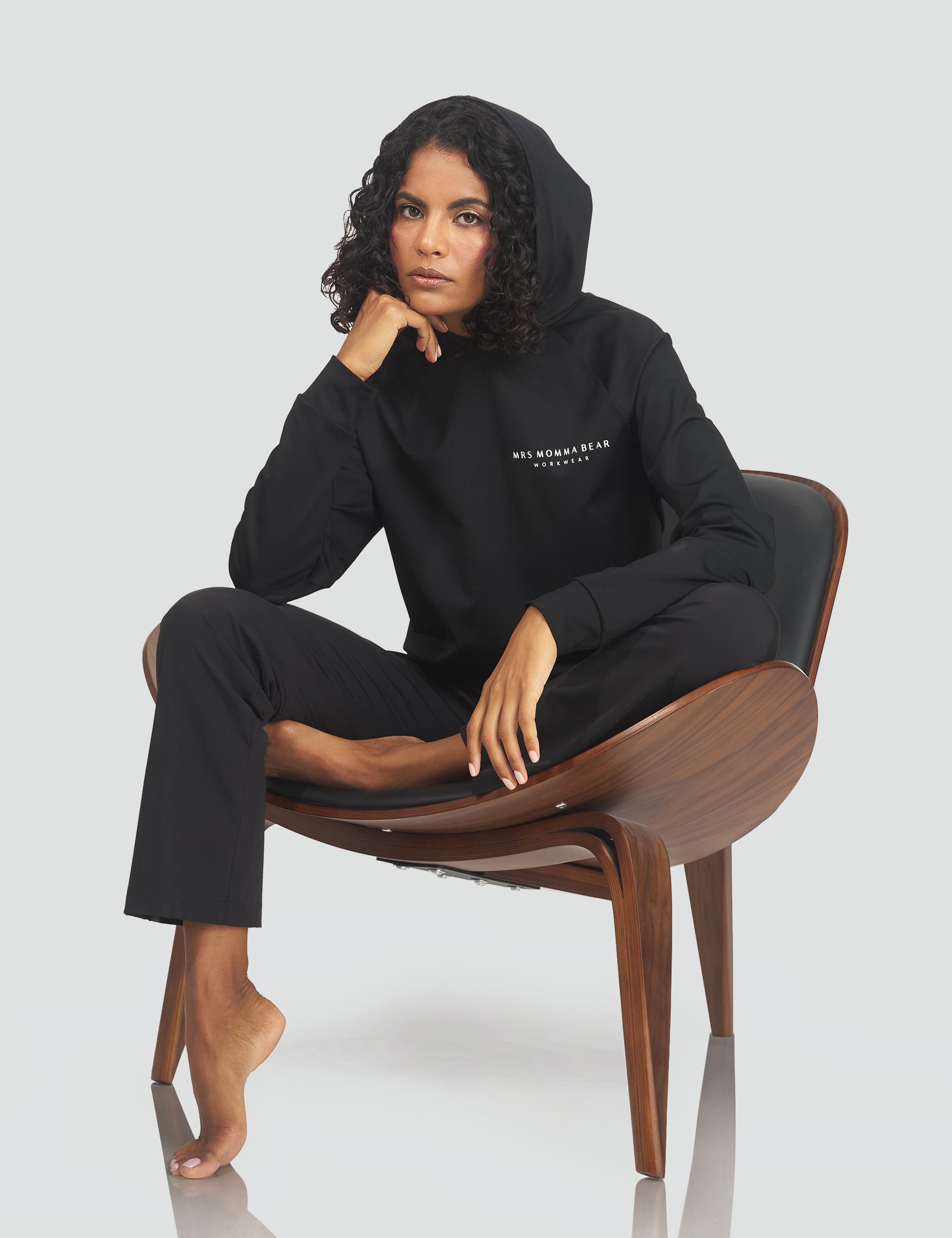 Woman in Original MMB Hoodie and pants sitting confidently on a wooden chair, showcasing modern athleisure wear.