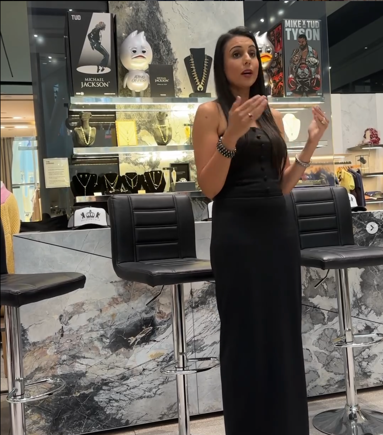 Woman in a black dress speaking confidently in a jewelry store, showcasing elegant pieces and decor.