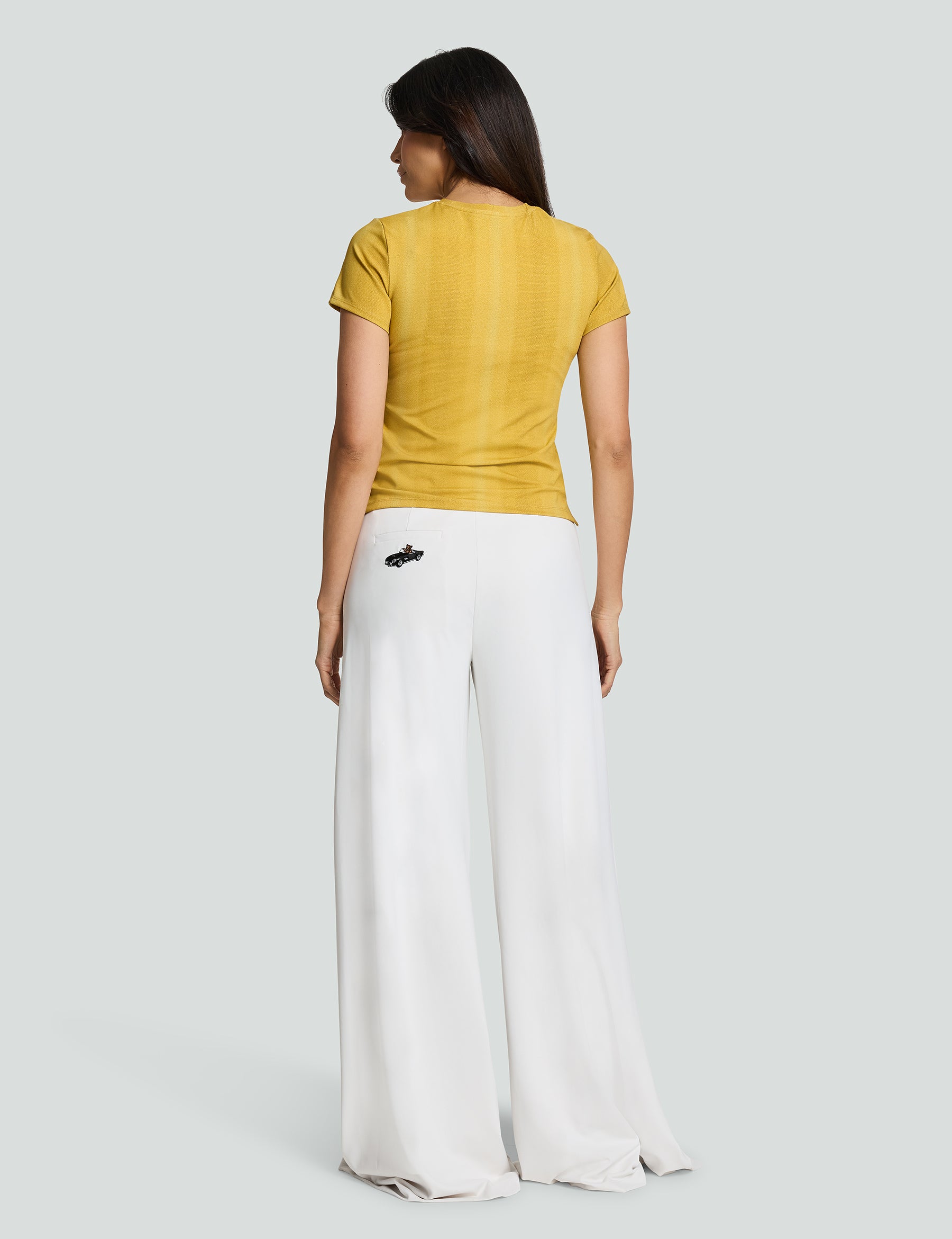 Back view of a woman wearing a gold Signature Luxe Tee paired with white wide-leg pants.