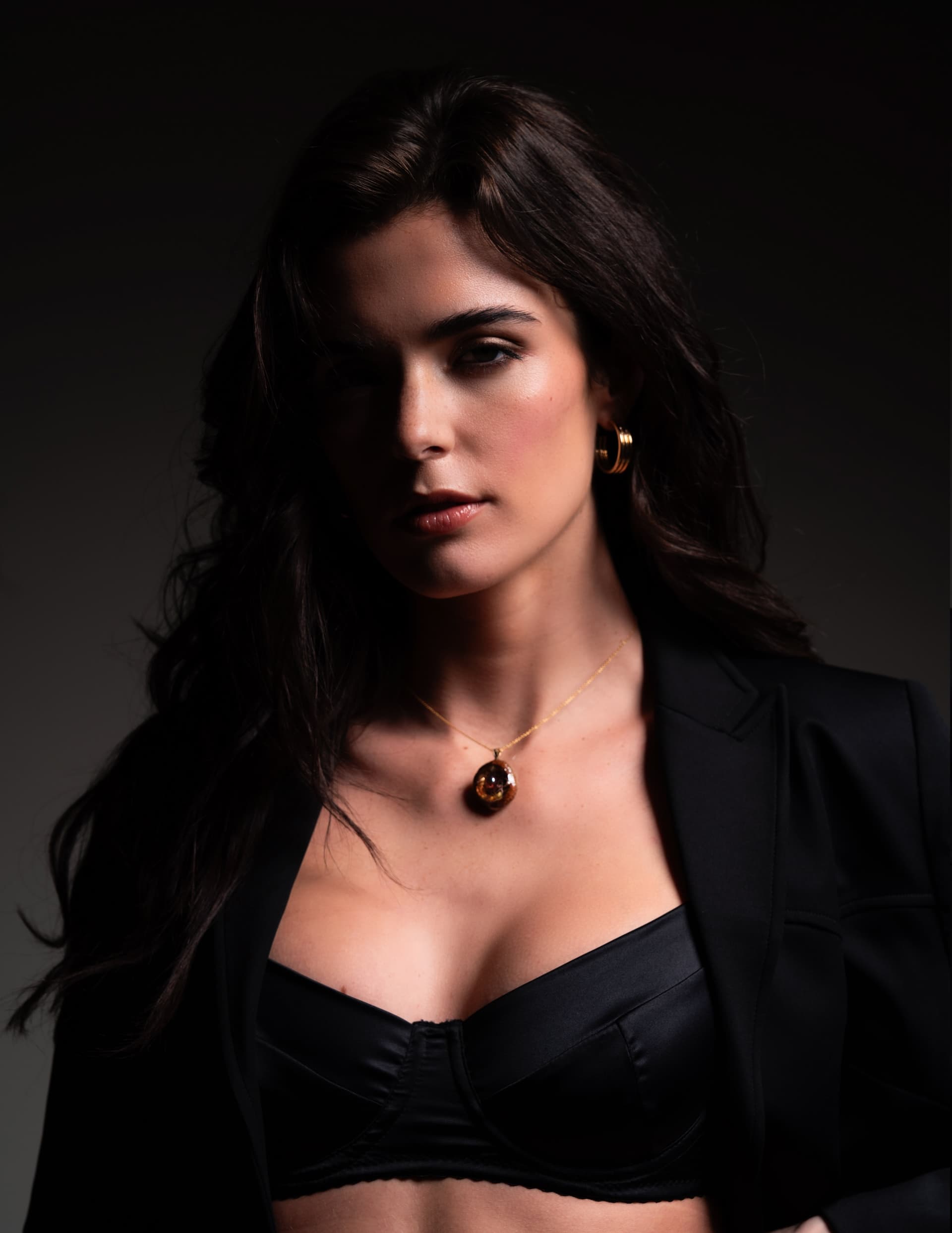 Model showcasing elegant jewelry, wearing a black bra and the You Hung the Moon blazer in dramatic lighting.