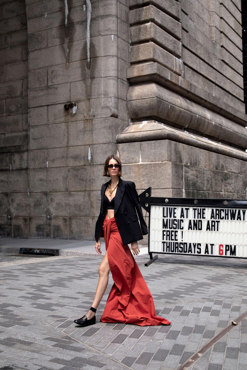 Fashionable woman in the Starry Night blazer and red skirt walks past outdoor event sign at The Archway, showcasing urban style.