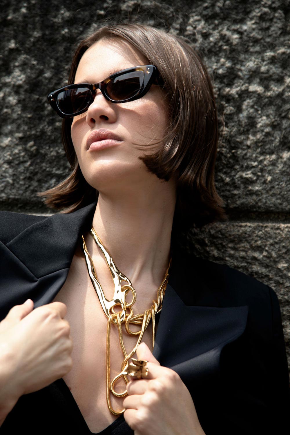 Model wearing black sunglasses and layered gold jewelry with the Starry Night Blazer against a textured stone backdrop.